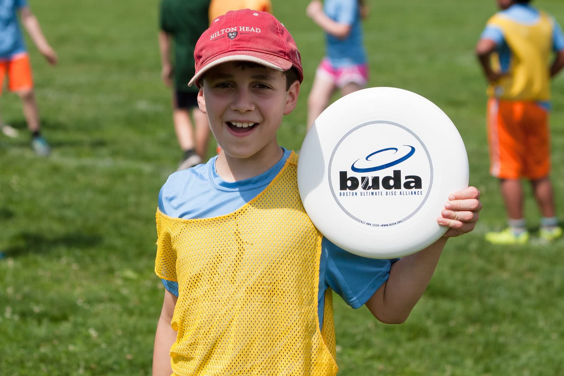 A young player in a yellow pennie grinning while holding up a BUDA disc